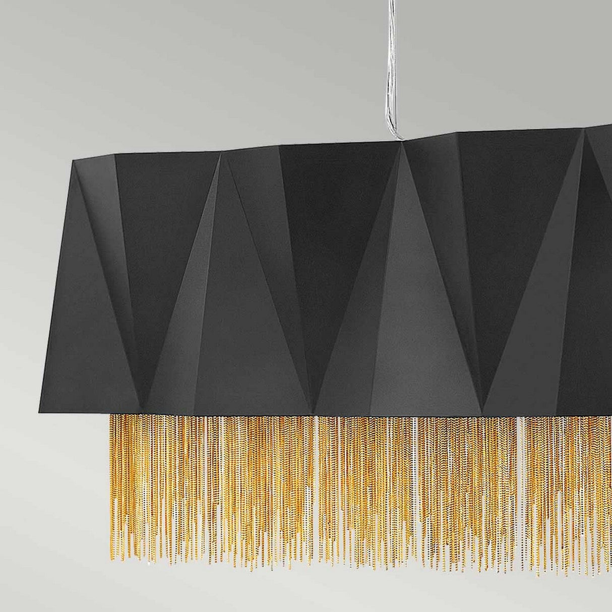 The Zuma 6 Light Linear Chandelier in Satin Black/Gold boasts a striking geometric design with a black faceted shade and elegant gold fringe details, enhancing any space with its modern lighting against a light gray background.