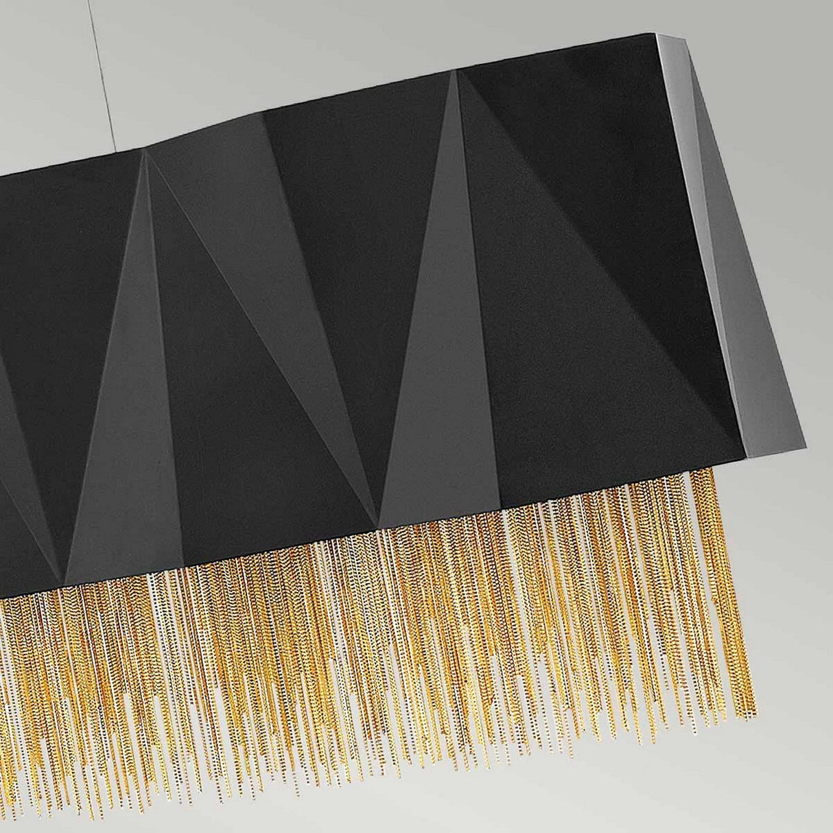 The Zuma 6 Light Linear Chandelier in Satin Black/Gold features a geometric design with a rectangular black pendant light, hanging gracefully from the ceiling. Gold chains drape from the bottom, accentuating its elegant and modern style against a simple, neutral-colored wall.