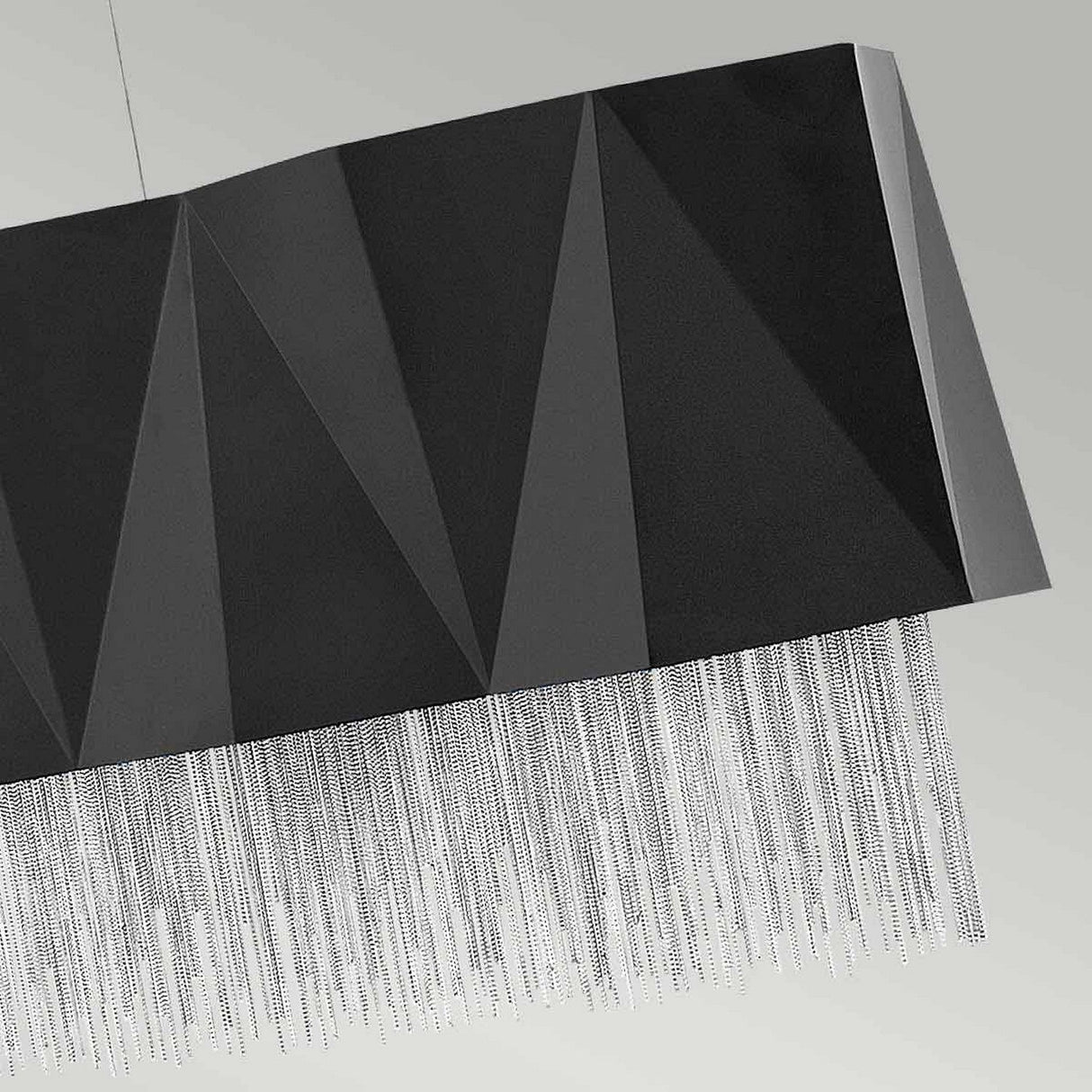 The Zuma 6 Light Linear Chandelier in Satin Black/Silver features a modern geometric design with a textured triangular pattern. It hangs elegantly against a light gray background, and its thin silver chains cascade down to create a fringe-like effect. This piece offers ample illumination and is ideal for enhancing contemporary interiors with its sleek style.