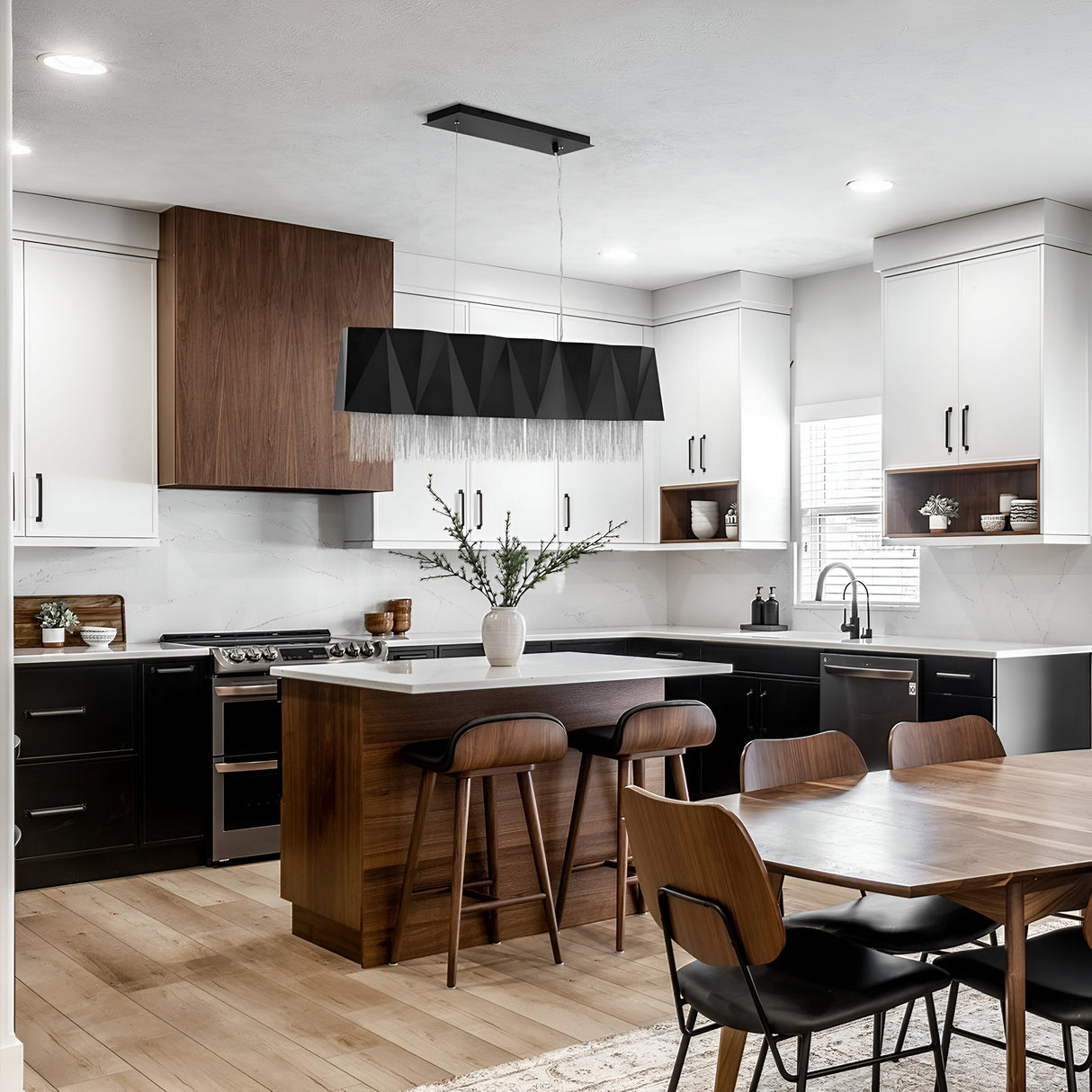 Modern kitchen with a central island showcasing the Zuma 6 Light Linear Chandelier in Satin Black/Silver, complemented by wooden barstools and a blend of dark wood and white cabinetry. A dining table with matching chairs is in the foreground, while ample natural light pours in through a window on the right.