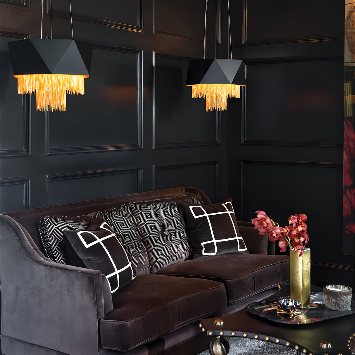 A stylish living room exudes contemporary elegance with its dark paneled walls and a plush velvet sofa accented by two geometric cushions. The Zuma 6 Light Chandelier in Satin Black/Gold bathes the room in a warm glow, while a metallic vase with red blooms graces the glossy black coffee table, adding a touch of satin black finish.