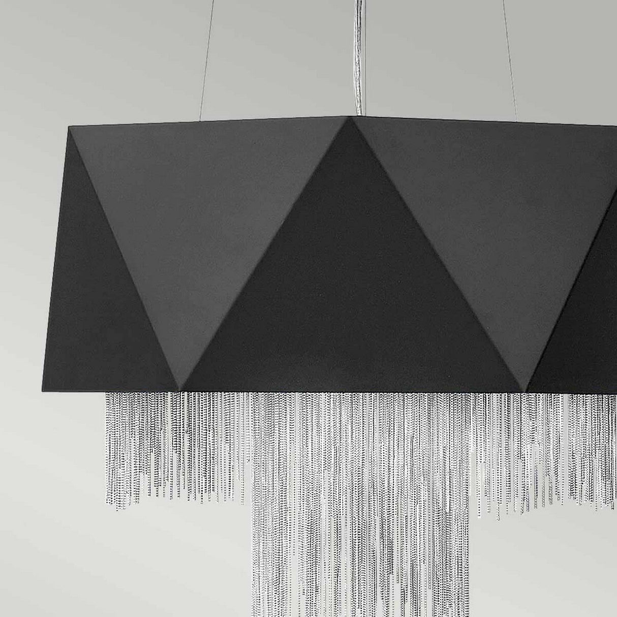 The Zuma 6 Light Chandelier in Satin Black/Silver showcases a modern geometric form with a textured, triangular design and silver chain-like fringes, elegantly set against a plain light gray background.