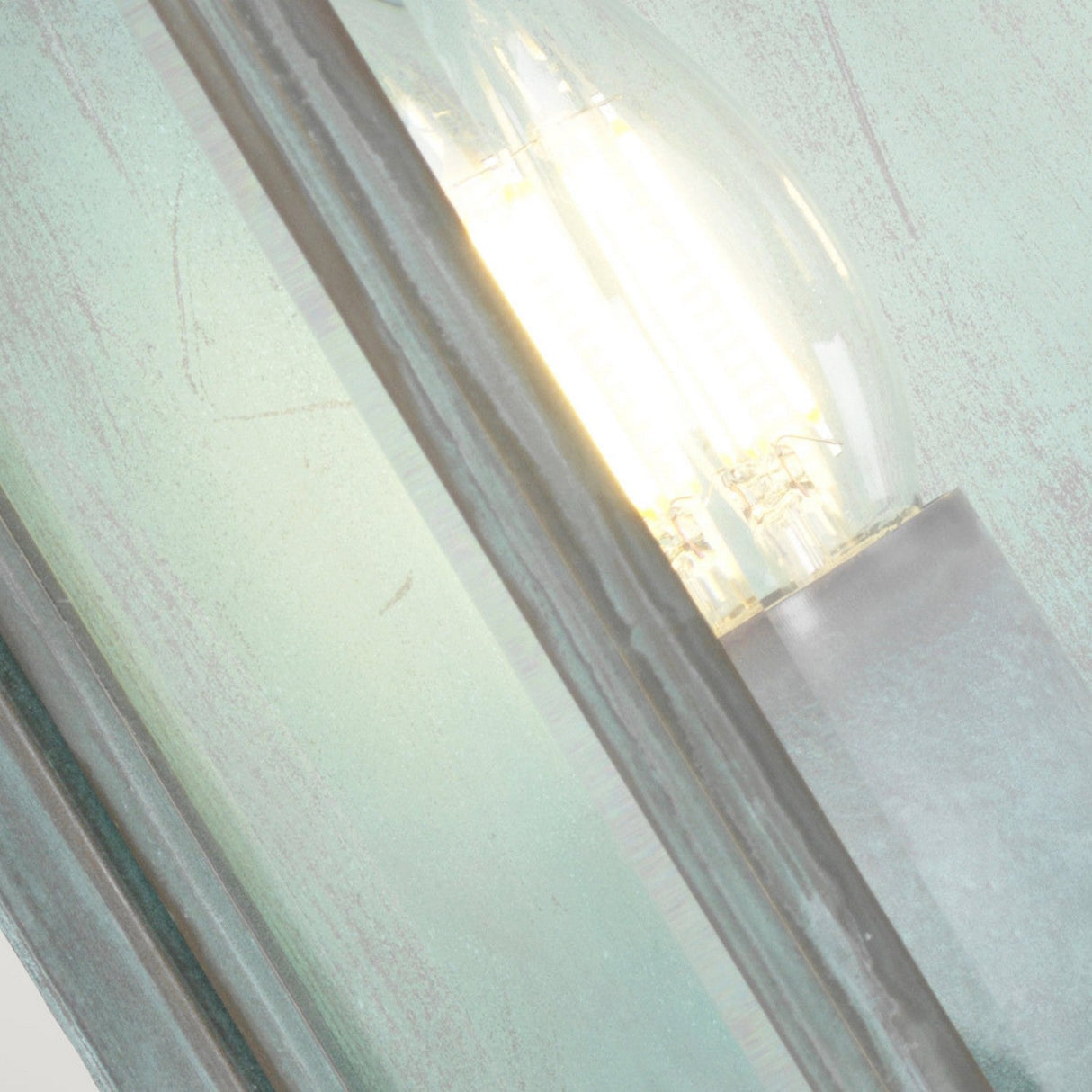 Close-up of a glowing Holborn 2 Light Outdoor Wall Light - Verdigris, encapsulated in frosted glass. The bulb emits warm light, enhanced by the verdigris finish that introduces a soft greenish tint. Crafted with solid brass construction, the design boasts a modern, minimalist aesthetic.