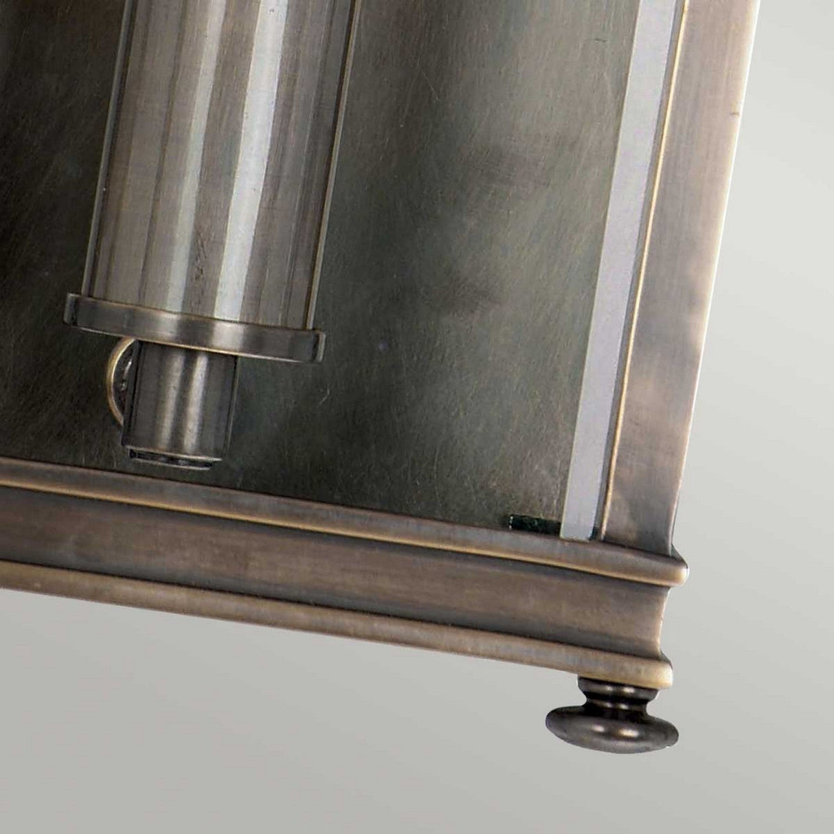 Close-up of the Holborn 1 Light Outdoor Small Wall Lantern in Dark Bronze, featuring a vintage metallic look with a cylindrical glass cover. This lantern, reminiscent of Georgian design outdoor lighting, showcases a metal frame with a tarnished finish and decorative rounded brass-like elements at the bottom corners.