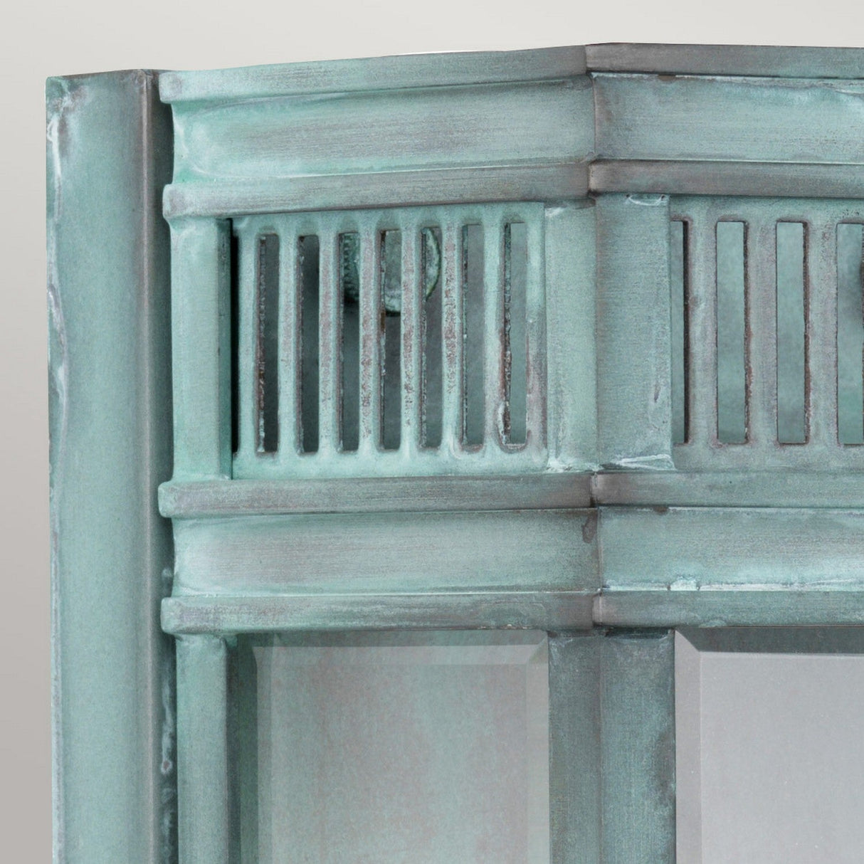 A close-up view showcases an aged, teal-tinted metal frame with decorative vertical slats and a verdigris finish. This structure, reminiscent of the Holborn 1 Light Outdoor Small Wall Lantern - Verdigris, features a weathered patina set against a plain, light gray background.