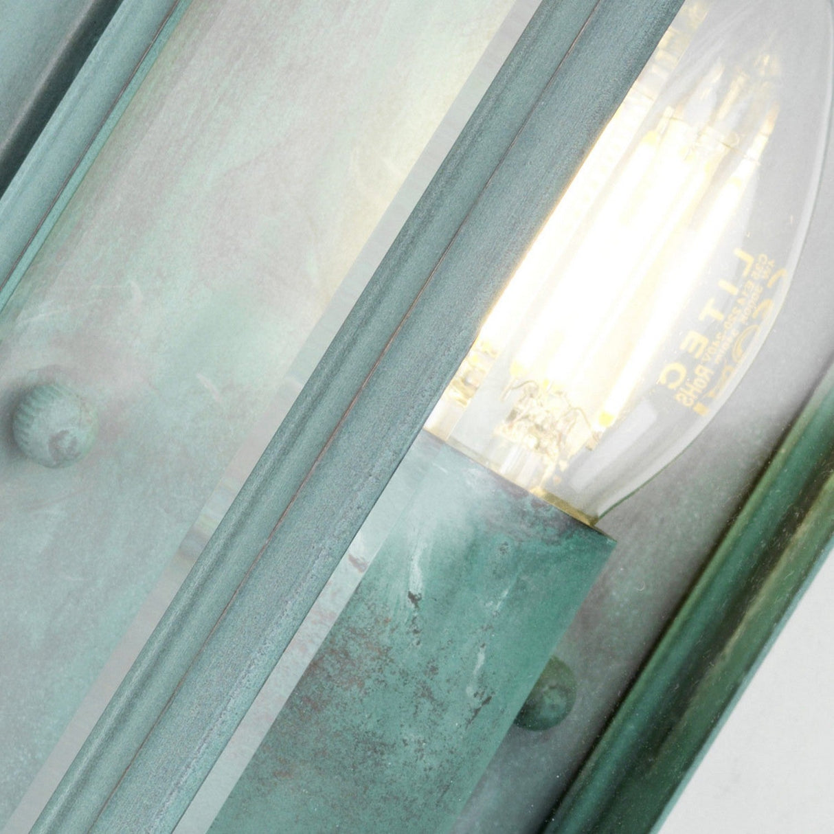 Close-up of the Holborn 1 Light Outdoor Small Wall Lantern - Verdigris, glowing warmly within a weathered, greenish metal frame. This outdoor wall light showcases clear glass panels and rustic patina, emphasizing the classic and industrial look of its Georgian design inspiration.