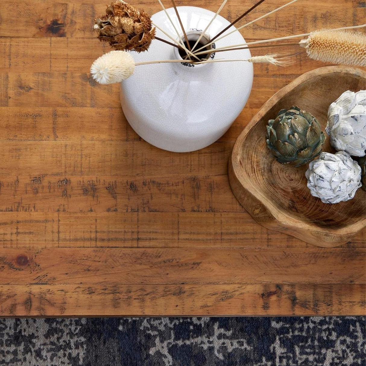 Hill Interiors antique bronze artichoke ornament in a wooden bowl with white pinecones, beside a white crackle-glaze vase of dried stems on rustic wood.