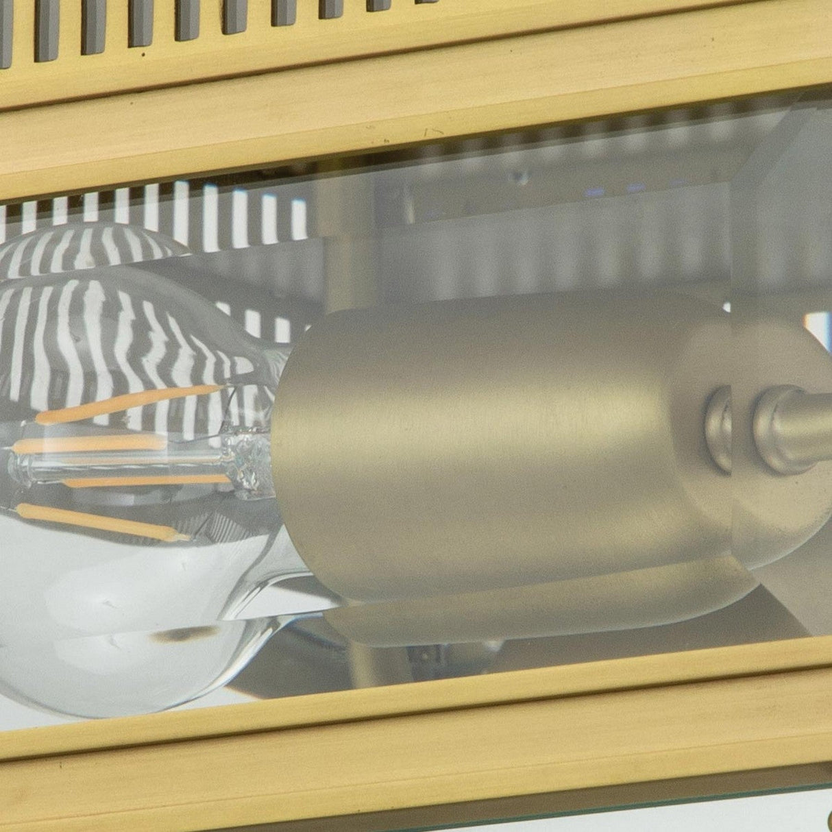 Close-up of the Holborn 2 Light Outdoor Flush fixture in brushed brass, showcasing an industrial-style design with a visible LED bulb encased in metal. The fixture combines reflective surfaces with a striped background pattern, making it ideal for both indoor and outdoor lighting settings.