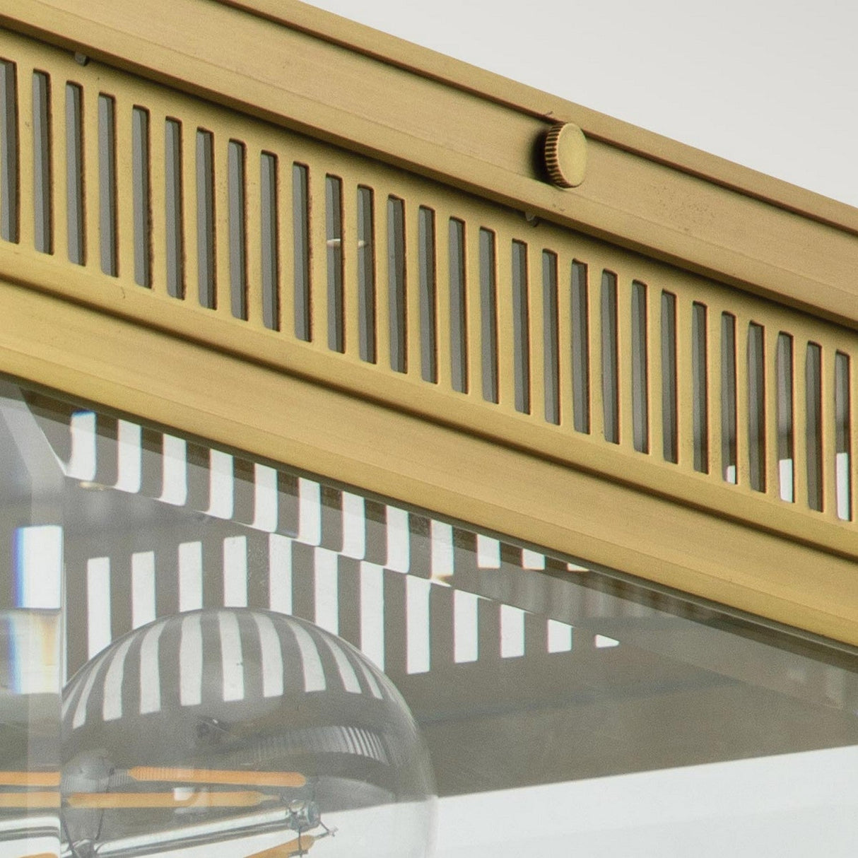 A close-up of the Holborn 2 Light Outdoor Flush in brushed brass and glass showcases its vertical slats. A partially visible light bulb inside reflects the slatted pattern on its surface, reminiscent of Georgian designs, capturing elegance against a neutral backdrop.