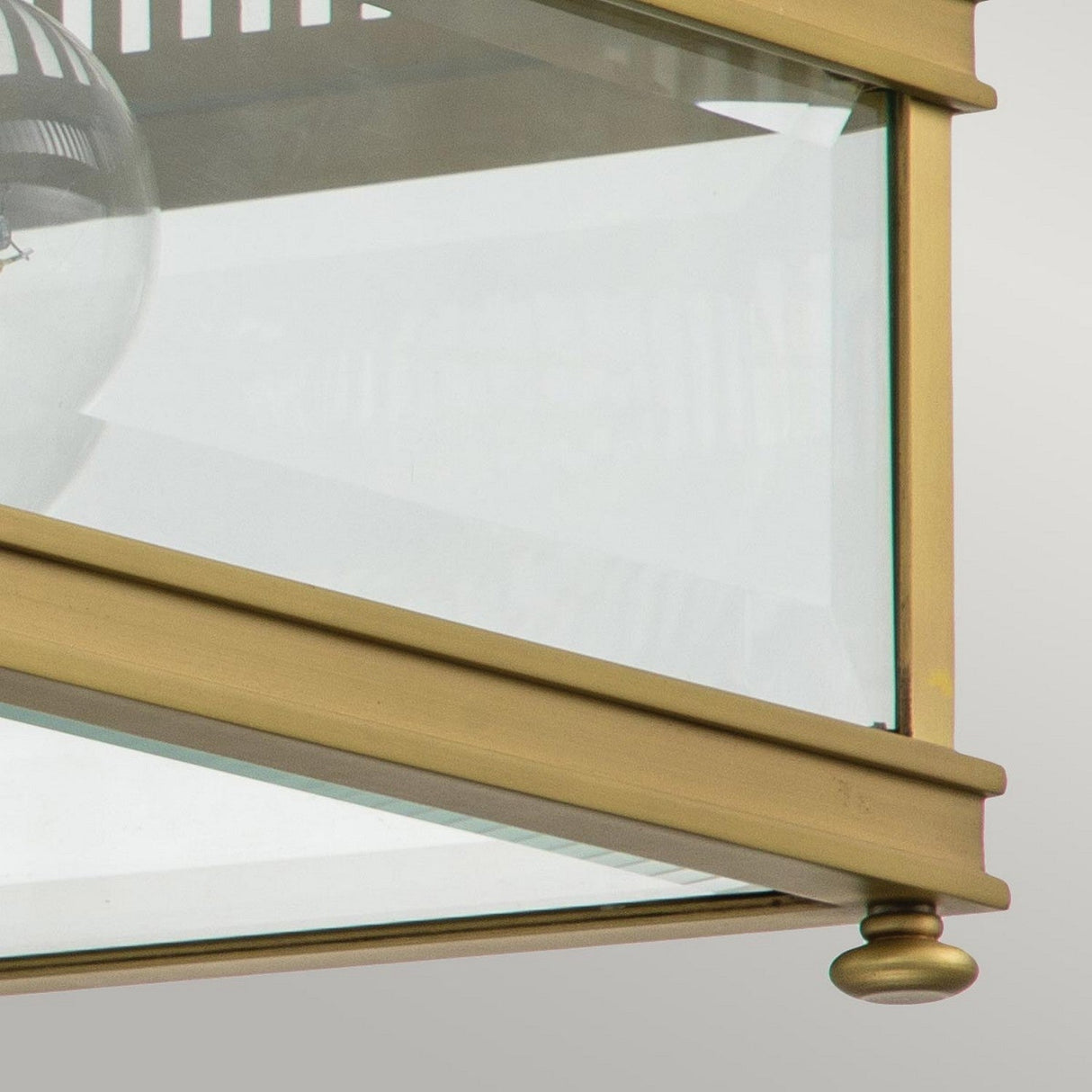 A detailed view of the Holborn 2 Light Outdoor Flush in brushed brass, inspired by Georgian designs. This fixture includes clear glass panels framed with brushed brass edges, highlighting a visible bulb inside. It is elegantly finished with a decorative knob at the bottom corner.