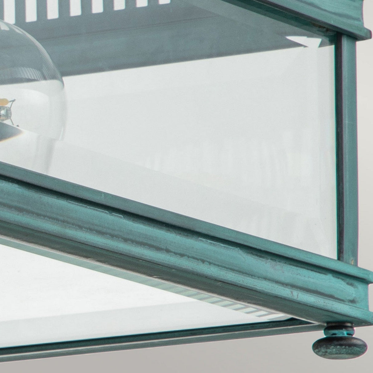 Introducing the Holborn 2 Light Outdoor Flush in Verdigris: This fixture features a vintage-inspired verdigris finish with a close-up view of its glass and metal design. The visible bulb is elegantly framed by transparent glass panels, while the textured metal surface offers an aged appearance, making it perfect for enhancing your outdoor lighting aesthetics.
