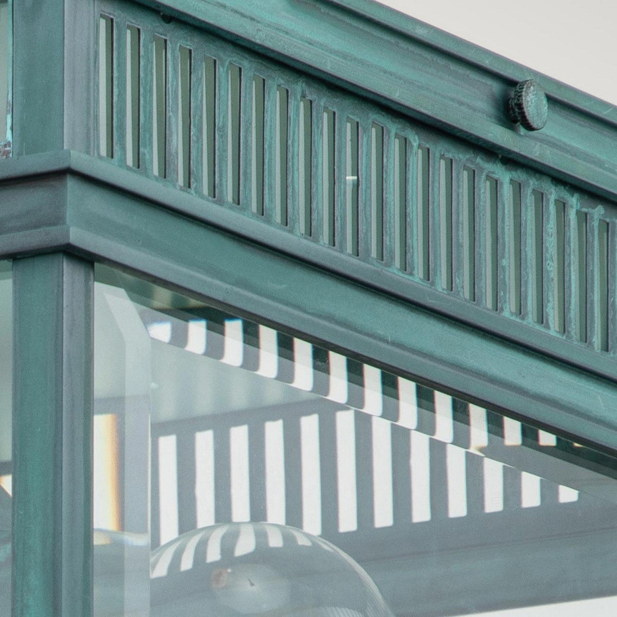 This close-up showcases the Holborn 2 Light Outdoor Flush - Verdigris, featuring a decorative metal and glass structure with a grid pattern. The verdigris finish imparts an aged green, vintage-inspired look to the metal, accented by vertical slits at the top, while the glass beautifully reflects light to produce a striped pattern.