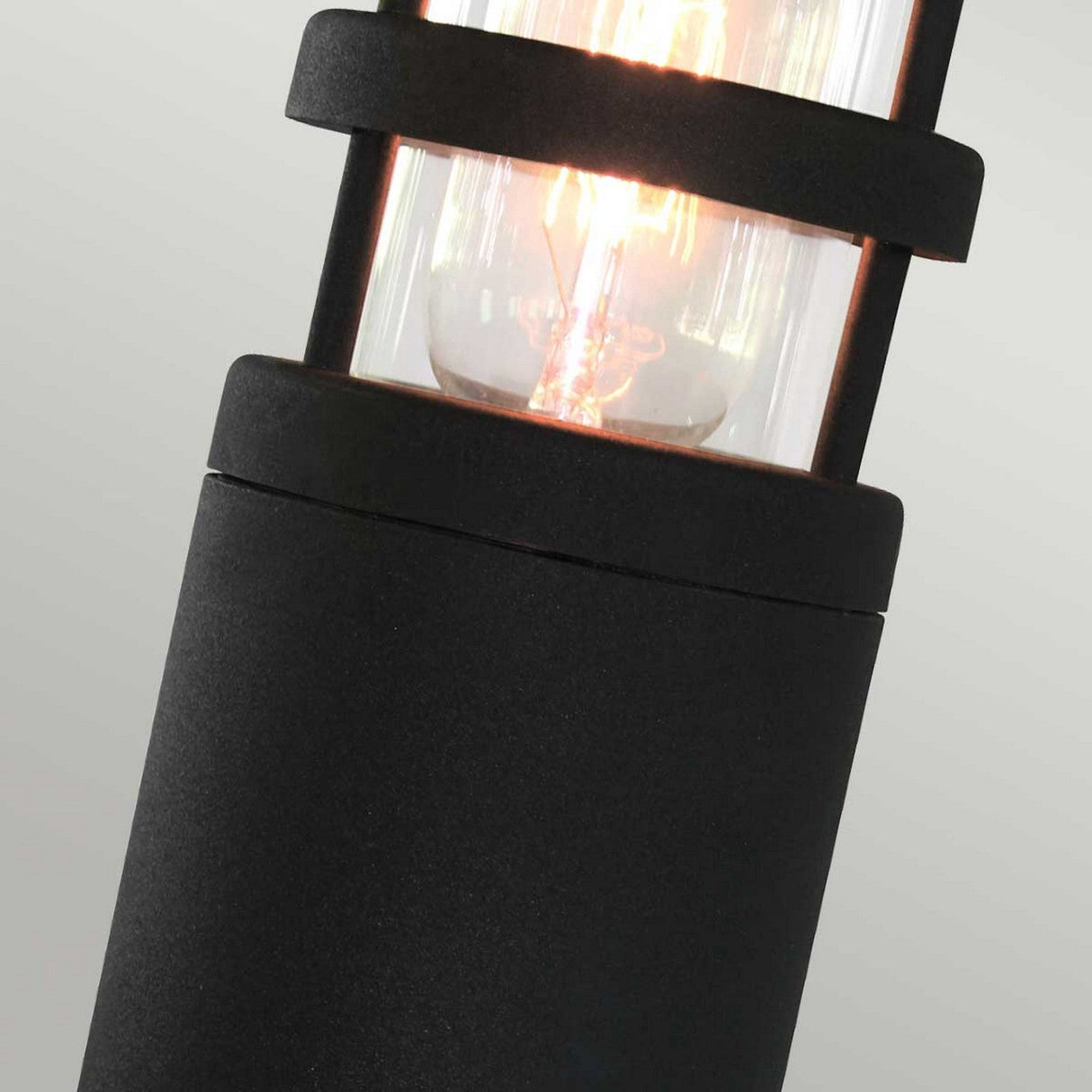 Close-up of the Hornbaek 1 Light Pedestal Light in black, showcasing its modern cylindrical shape with a visible glowing filament encased in clear glass. This lamp exhibits Scandinavian design elements with a sleek matte finish and strong geometric lines typical of contemporary pedestal lights.