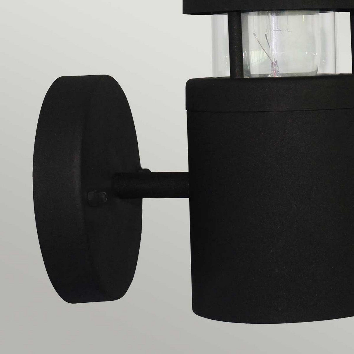 The Hornbaek 1 Light Outdoor Wall Light in black features a sleek, cylindrical design with a transparent upper section and is mounted on a circular base. Its Scandinavian simplicity exudes modern charm against a light gray backdrop, while its durable construction guarantees long-lasting elegance for your exterior lighting needs.