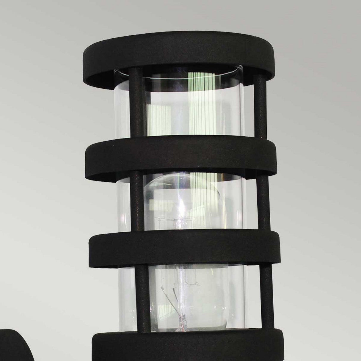 Close-up of the Hornbaek 1 Light Outdoor Wall Light in black, featuring a sleek cylindrical glass shade encased in a black metal frame that highlights Scandinavian simplicity. The design includes vertical bars for a modern, contemporary look and offers durable outdoor performance against a plain light gray background.