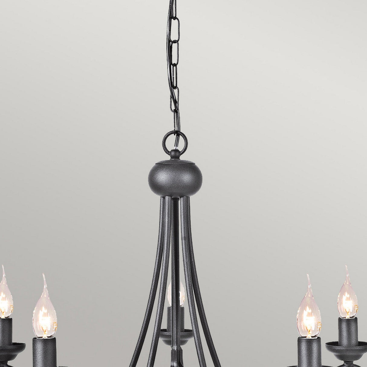 A close-up of the Harlech 5 Light Chandelier - Black features lit candle-style bulbs, with a black metal chain that enhances its rustic charm against a plain, light-colored wall. This chandelier exudes an elegant yet timeless ambiance.