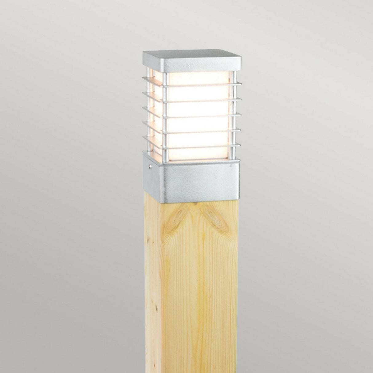 The Halmstad Large Bollard Natural Wood - Silver adds a modern touch to any outdoor area. It boasts a natural wood finish combined with a sleek, rectangular stainless steel fixture. Vertical bars highlight the glowing bulb against a neutral backdrop.