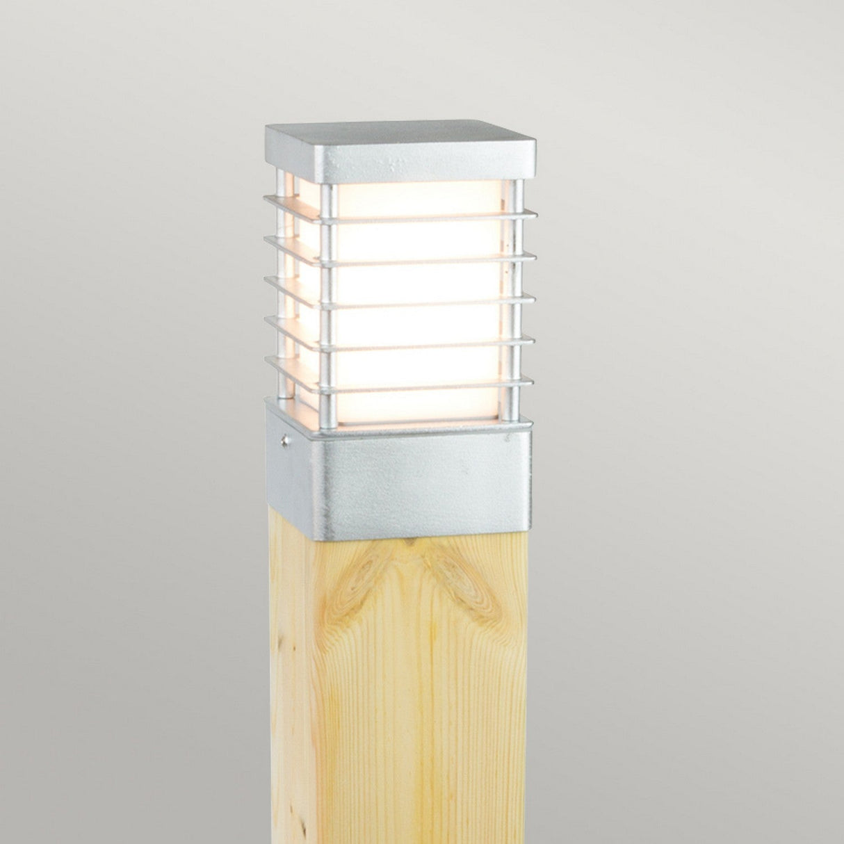 The Halmstad Medium Bollard Natural Wood - Silver is a contemporary outdoor post light designed with a natural wood base and a square metallic fixture. Its medium-sized bollard features a grid design that casts a gentle, diffused glow, making it ideal for illuminating garden pathways or driveways. The backdrop is simple and light gray.