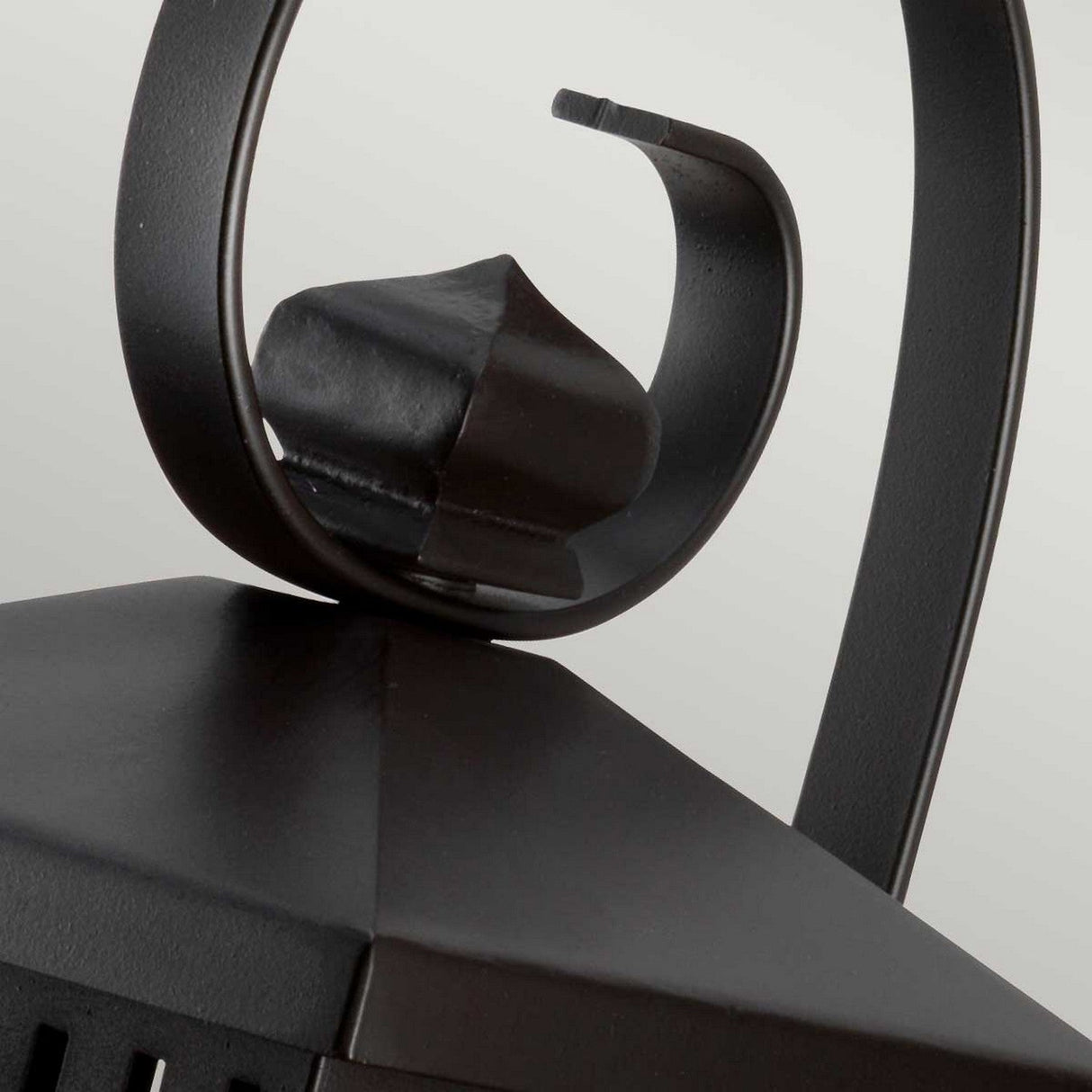 Detailed view of the Hythe 1 Light Outdoor Wall Light, showcasing its pyramid-shaped top. This black metal fixture has a curled handle and sleek, clean lines with an elegant old bronze finish, set against a neutral backdrop.