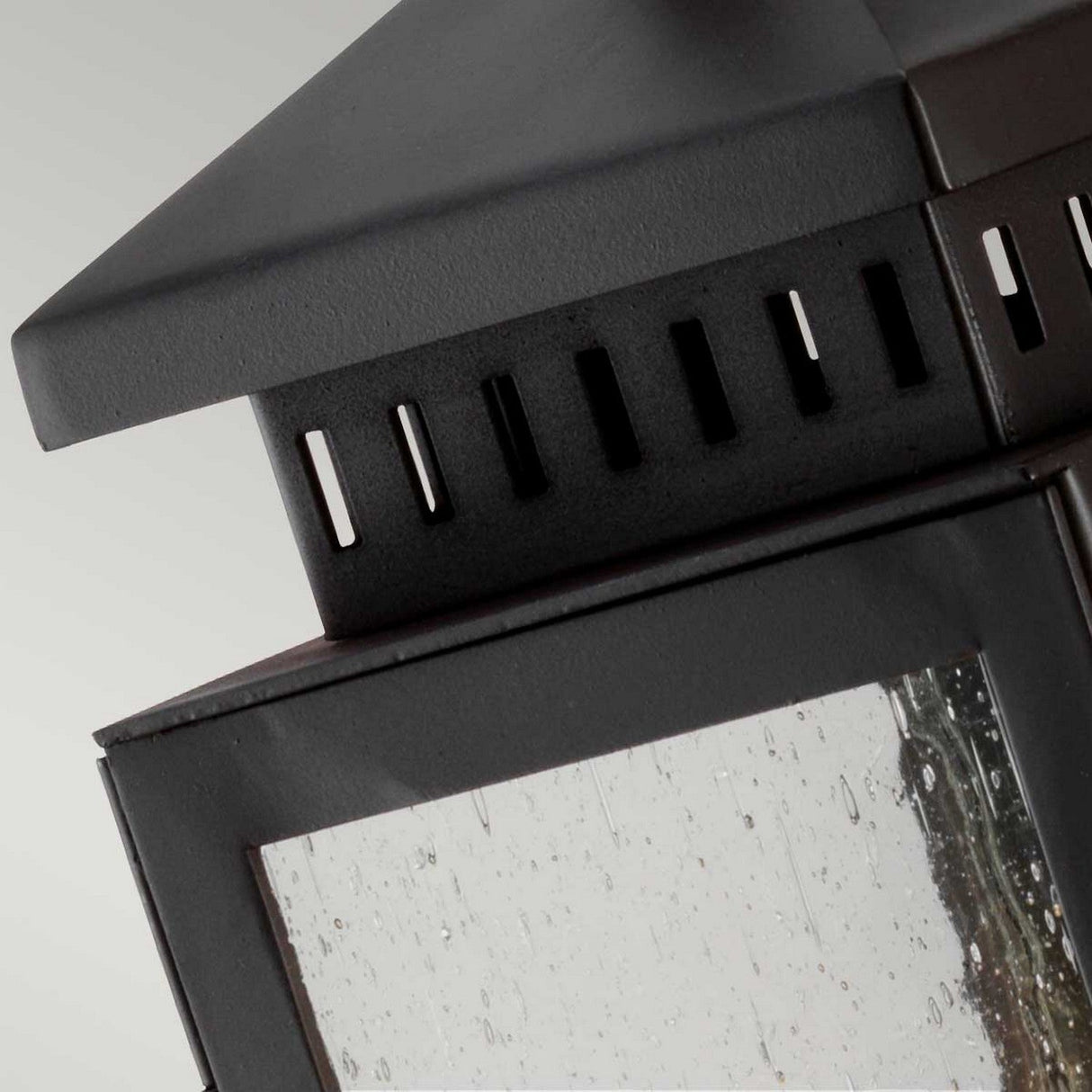 A close-up shot showcases the upper section of the Hythe 1 Light Outdoor Wall Light, crafted from black metal with a vented design and glass panel. The subtly speckled glass looks wet, accentuating its contemporary aesthetic. This fixture features a pyramid-shaped top and is finished in old bronze, bringing elegance to any setting.