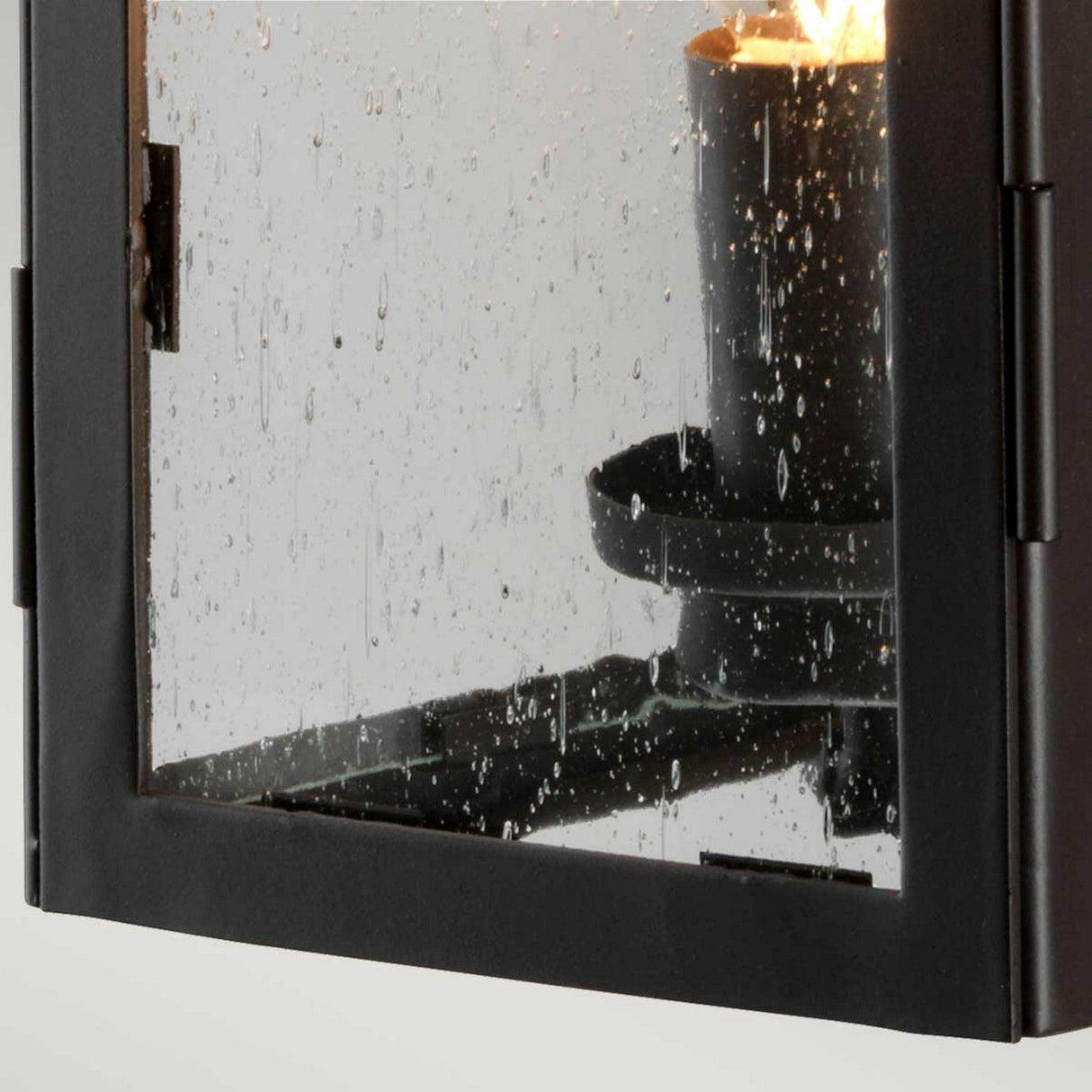 Close-up of a wet glass panel set in an Old Bronze metal frame from the Hythe 1 Light Outdoor Wall Light, showing drops of water forming a blurry texture on the surface. The wall light is visible behind the glass, creating a warm glow through the droplets.