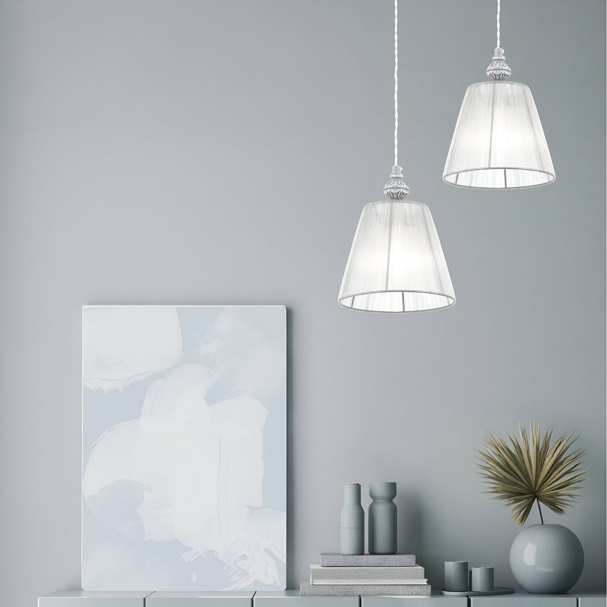 Two elegant pendant lights, including the Monsoon Pendant Light with an antique gray organza shade, brighten a minimalist space. Beneath them, an abstract painting, stacked books, decorative bottles, and a spiky gold sculpture are neatly displayed on a pale console.