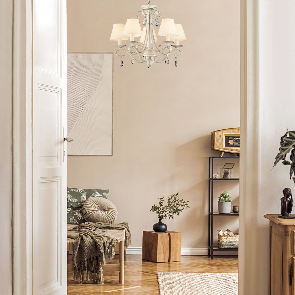 A cozy room with a white door reveals a seating area featuring a patterned sofa, round cushion, and throw. The Brionia 5 Light Chandelier in Beige & Gold hangs elegantly from the ceiling. Nearby, a shelf holds books and an old radio, while a small plant rests on a wooden cube.