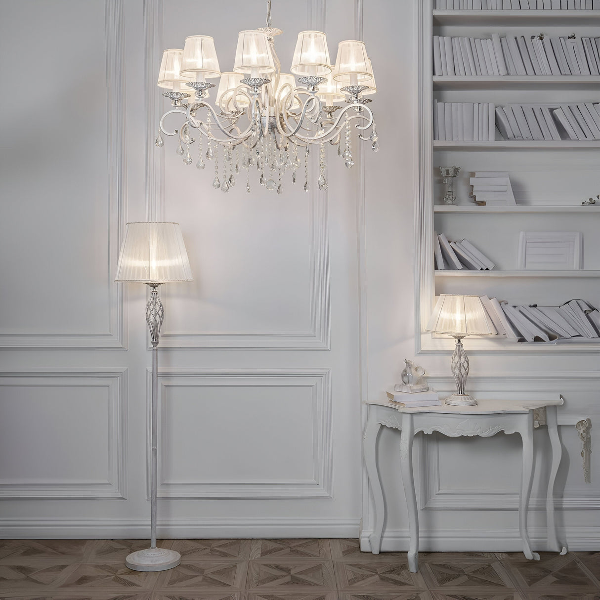An elegant white room features a chandelier with multiple lampshades, a floor lamp, and a small Grace Table Lamp - White & Gold on an ornate table. White shelves filled with books line the wall, adding sophistication highlighted by touches of white and gold.