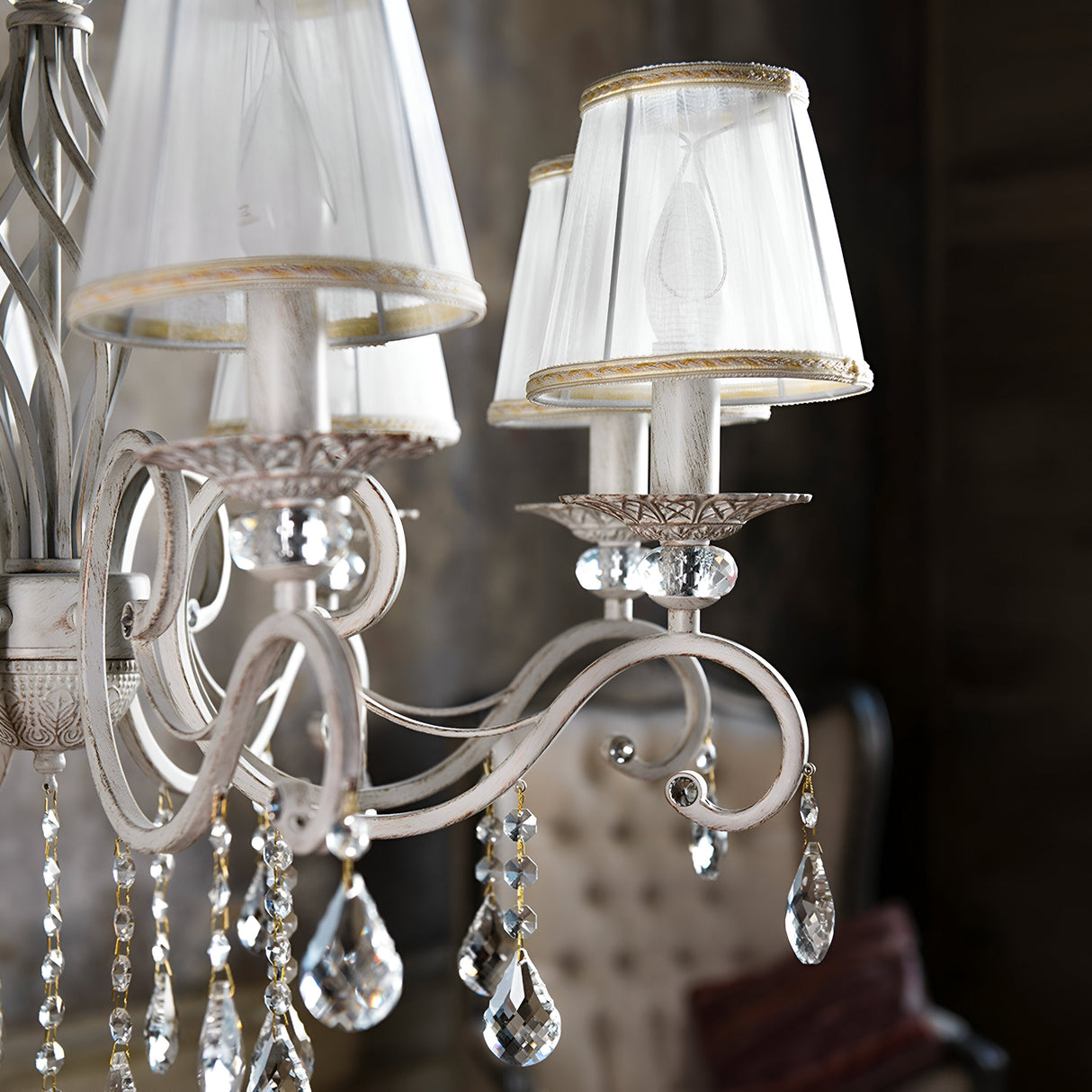 A close-up of the Grace 8 Light Chandelier - White showcases its cream organza lampshades and crystal pendants on curved metallic arms. The softly blurred background features a tufted chair, enhancing a vintage and luxurious atmosphere.