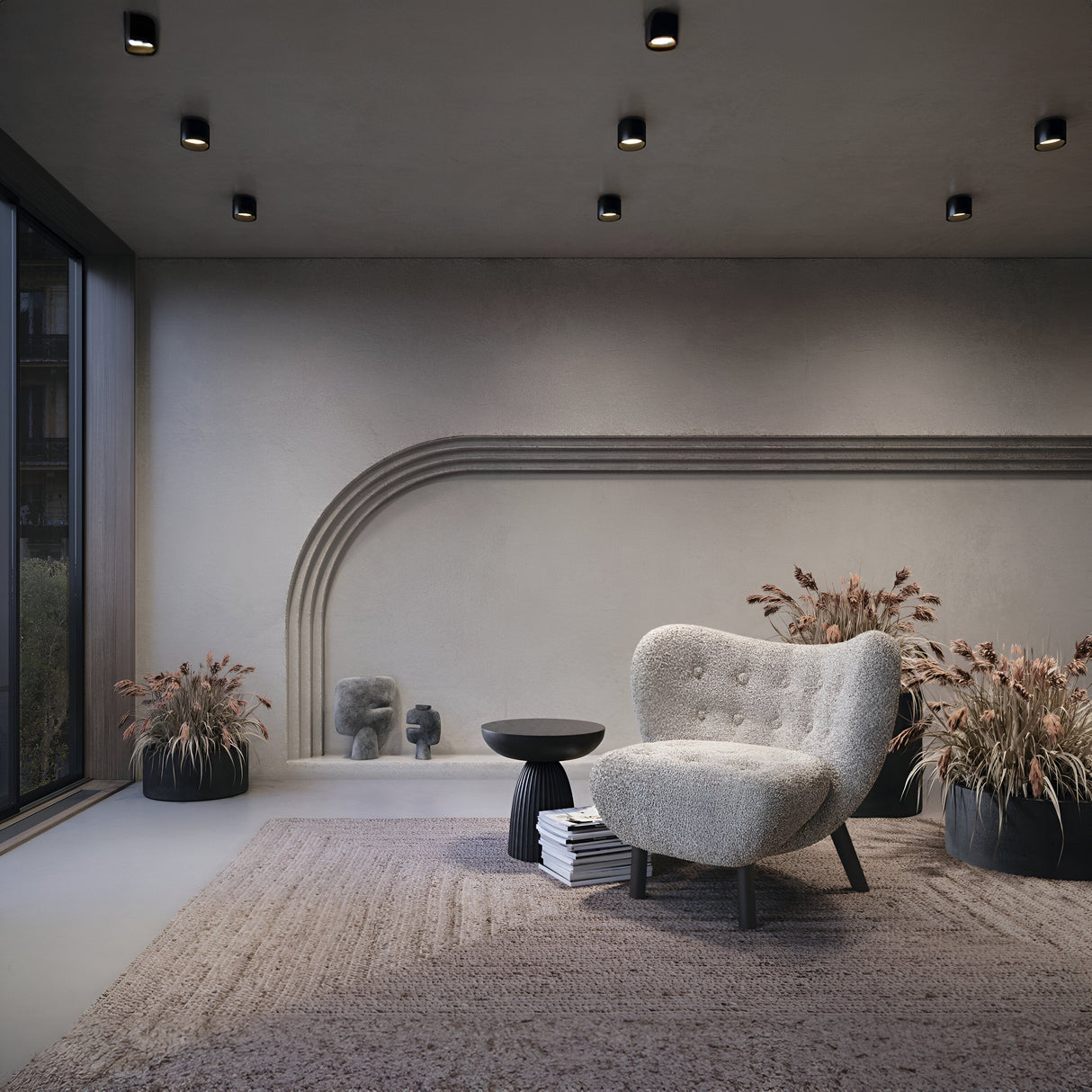 A modern living room features a curved gray armchair and small black round table on a textured rug. Decorative plants in black pots line minimally arched walls. The ambiance is enhanced by the Planet LED Small Round Surface Downlight 3000K - Black, while a large window provides natural light.