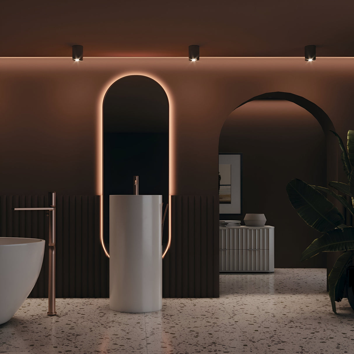 A modern bathroom features a sleek pedestal sink with an oval backlit mirror. The Slim Round Flat Surface Mounted Downlight in black enhances the warm lighting, highlighting terrazzo floors and minimalist design. A white tub is partially visible, complemented by a large plant.