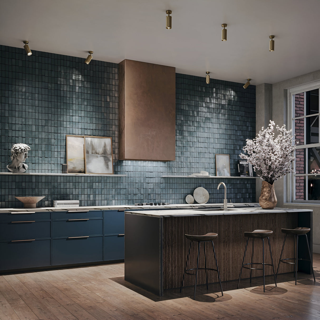 The modern kitchen showcases a dark blue tiled wall and a central island with brown paneling and a marble countertop. Three black swivel stools line the island, while minimalist decor enhances the space. A large vase with cherry blossoms sits by the window, underneath a Focus Design Single Spotlight in Matte Gold.