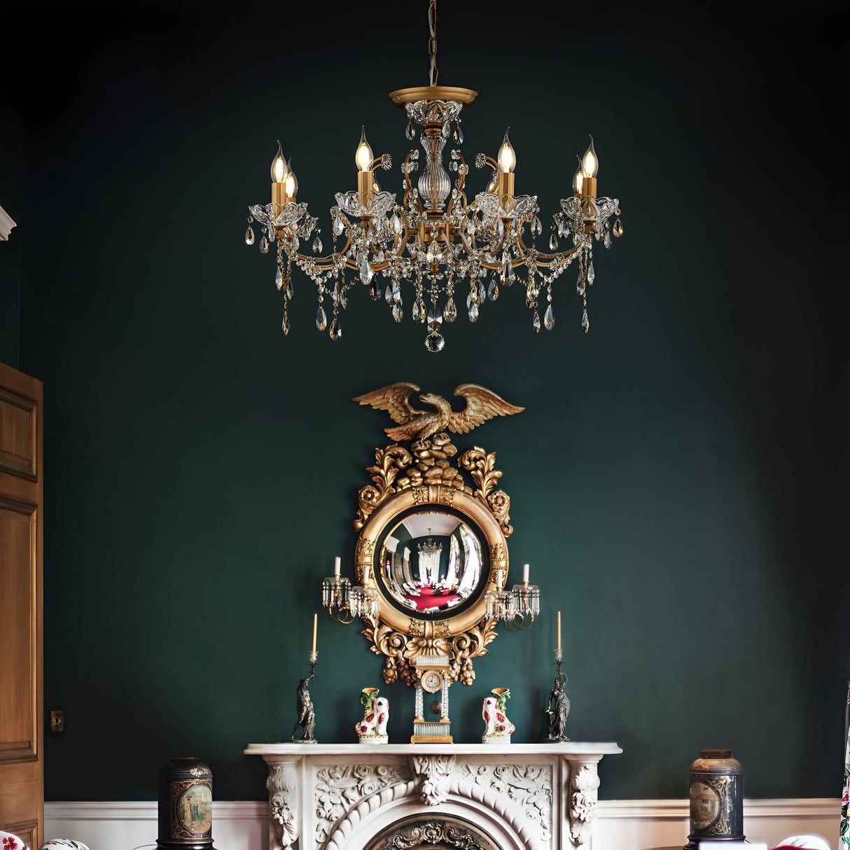 A luxurious room with a dark green wall features the Sevilla 8 Light Chandelier in gold, illuminating an antique mirror topped with an eagle and an elegant fireplace adorned with vases, figurines, and decorative crystals.