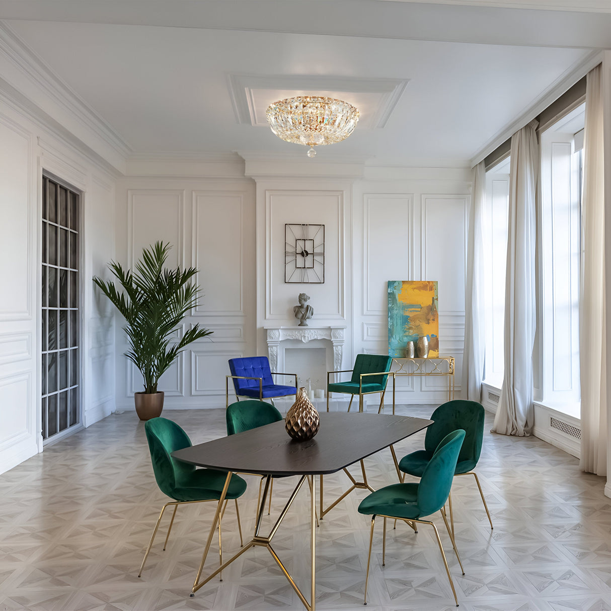 A modern dining room showcases a dark table with green and blue velvet chairs, a lush plant, and an abstract painting on white walls. The Basfor 3 Light Crystal Chandelier in gold adds luxurious lighting while natural light floods through large windows.
