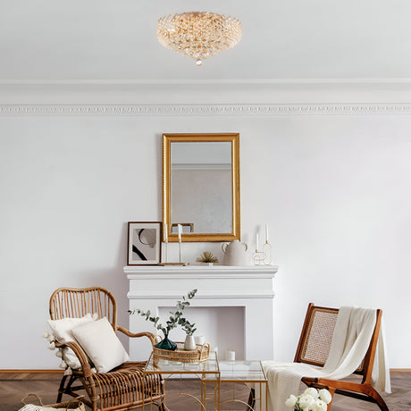 A cozy living room showcases a white fireplace, a golden-framed mirror, and two wooden chairs with cushions flanking a small glass-top table. The room is adorned with plants, candles, and the Karolina 9 Light Chandelier in gold, featuring sparkling crystal pendants.