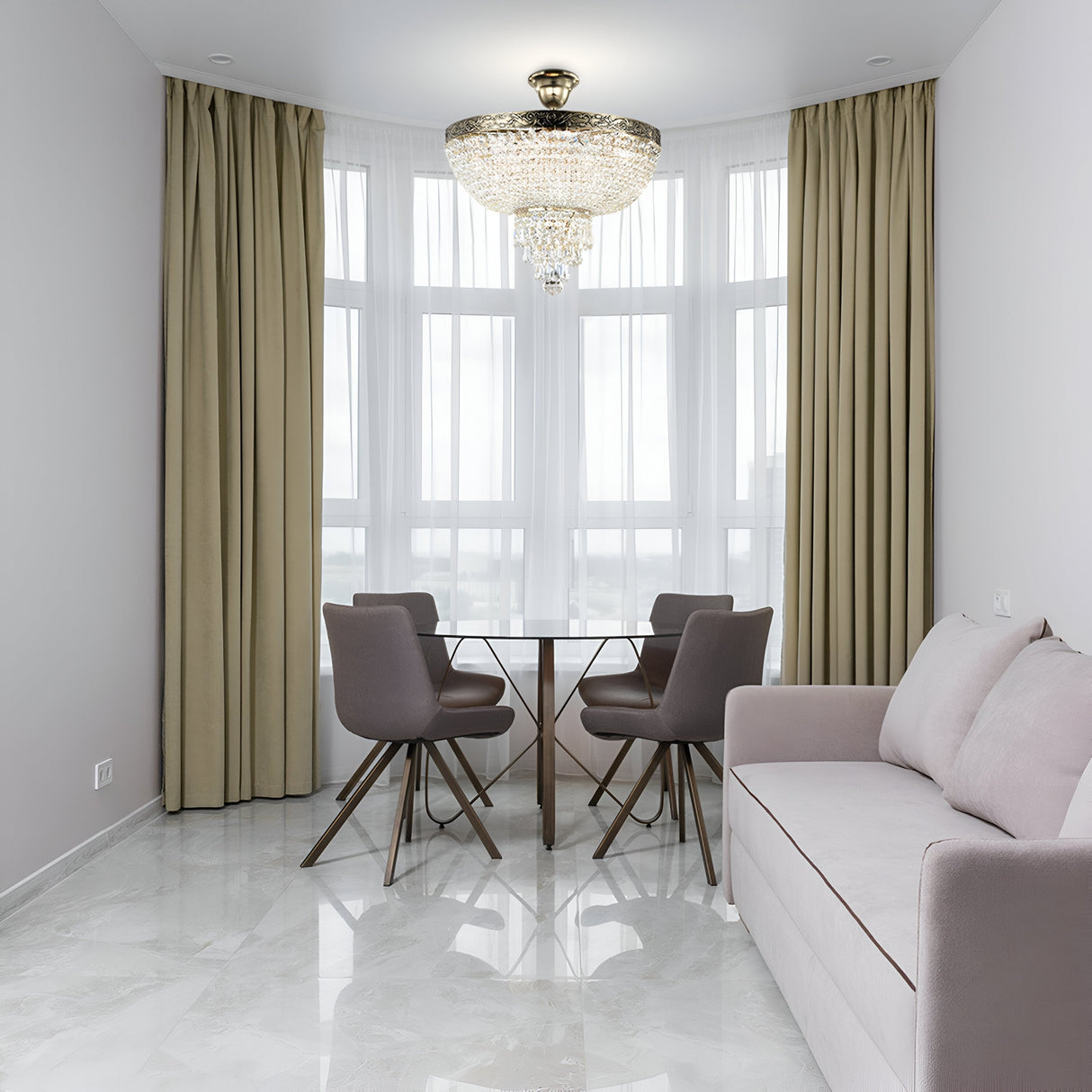 A modern dining area features a round glass table with four gray chairs by a window with beige curtains. A beige sofa sits on the right. Overhead, the Palace 5 Light Crystal Chandelier in gold casts an elegant glow. The room is adorned with white walls and a glossy tiled floor.