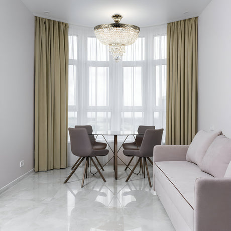 A modern dining area features a round glass table with four gray chairs by a window with beige curtains. A beige sofa sits on the right. Overhead, the Palace 5 Light Crystal Chandelier in gold casts an elegant glow. The room is adorned with white walls and a glossy tiled floor.