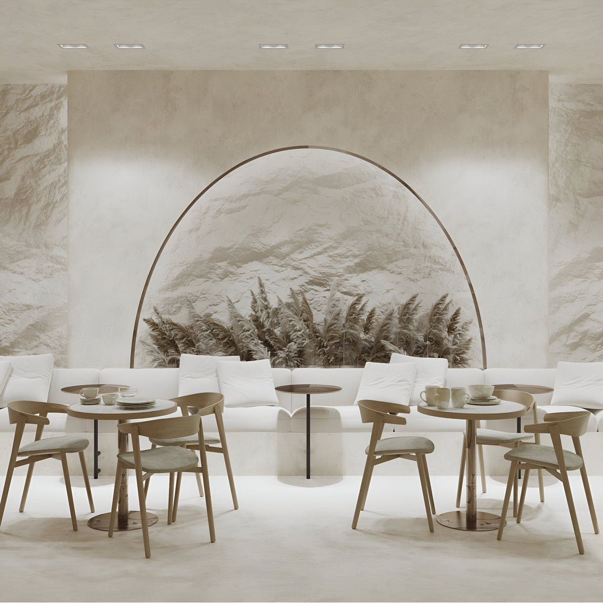 A minimalist cafe interior with a neutral color scheme features a mountain mural. Round wooden tables and modern chairs accompany a long cushioned bench with white pillows, softly illuminated by Atom Twin Tilt Rectangular Plaster-in Recessed Downlights, adding subtle directional lighting.