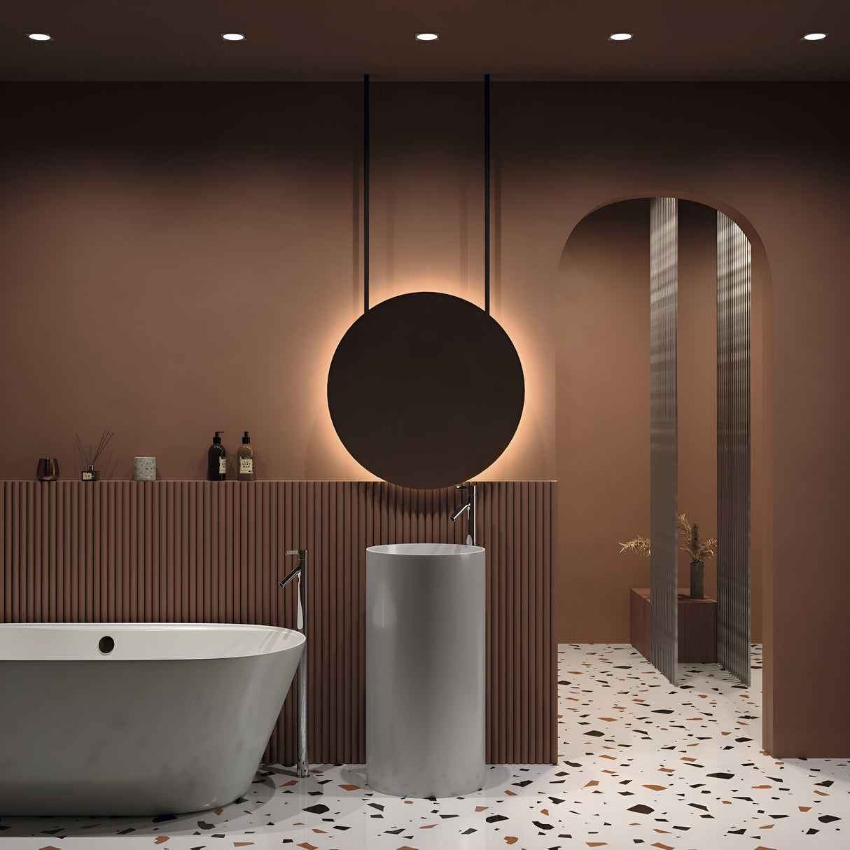 A modern bathroom with brown walls features a round backlit mirror above a white cylindrical sink. A freestanding bathtub sits on the left, and an arched doorway adorns the right. The terrazzo floor and minimalist design are highlighted by a Zoom Single Fixed Square Recessed Downlight - White.