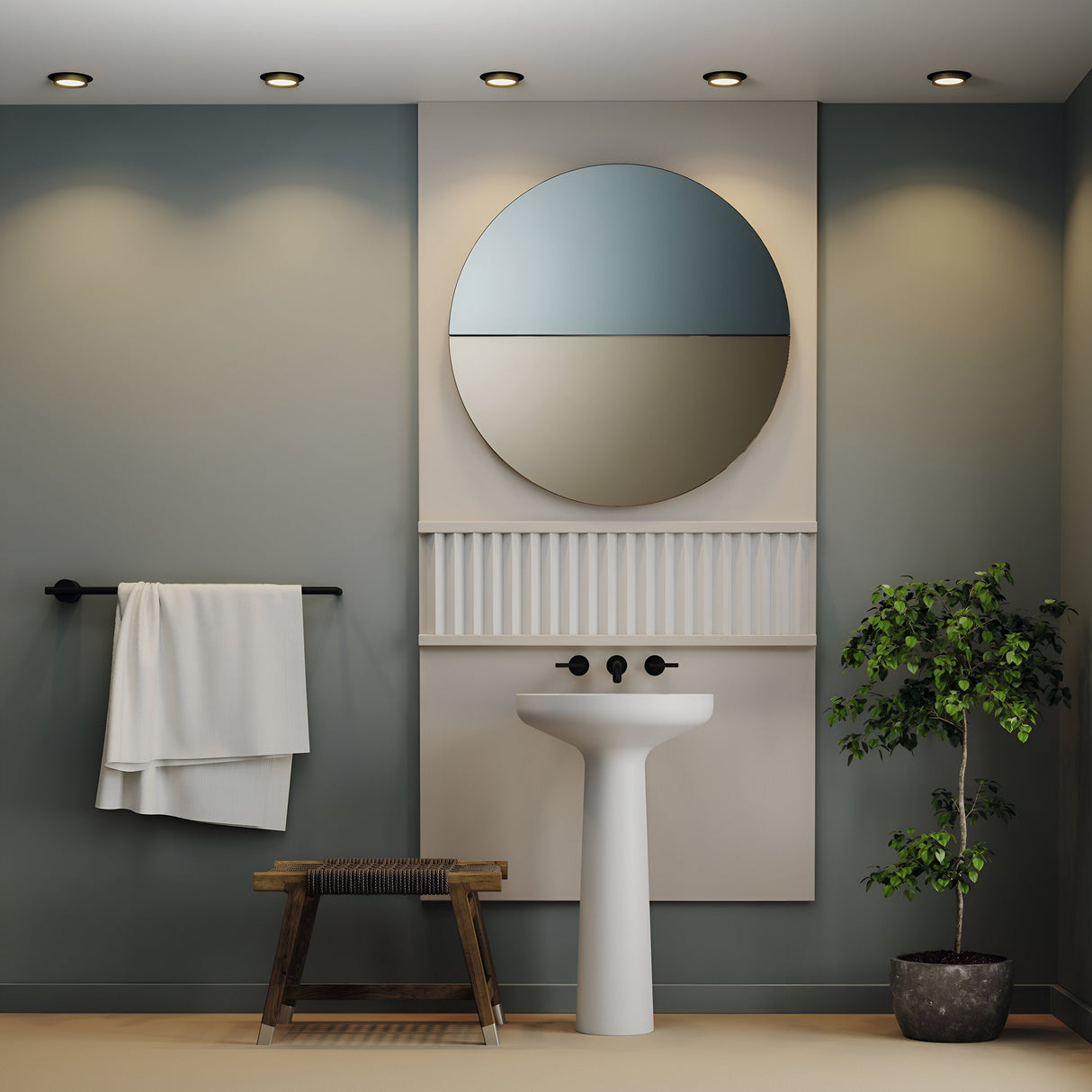A modern bathroom boasts a round mirror above a white pedestal sink, with a towel on a black rack beside a wooden bench. A potted plant stands to the right, lit by energy-efficient Hoop Single Fixed Round Recessed Downlights in Black & Gold. The walls are painted green-gray.
