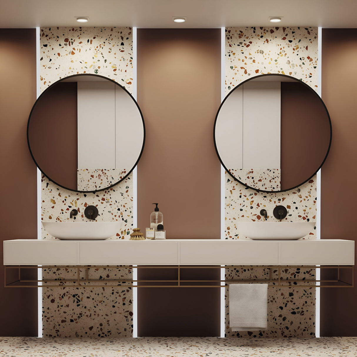 Modern interior design features a contemporary bathroom with two round mirrors above white sinks on a terrazzo-patterned wall. A white countertop holds soap dispensers, while a towel hangs below. The Hoop Single Fixed Square Recessed Downlight - White casts warm lighting, highlighting the elegant brown and white decor.