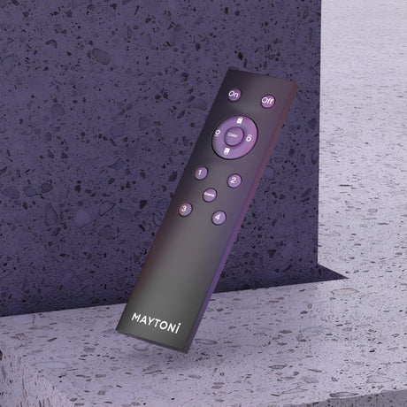 The Exility Smart Lighting Remote Control - Black, branded with MAYTONI, leans against a speckled stone surface. It features buttons for power, color, brightness, and navigation in a circular layout. Sync it with the Maytoni Smart app to enhance functionality.