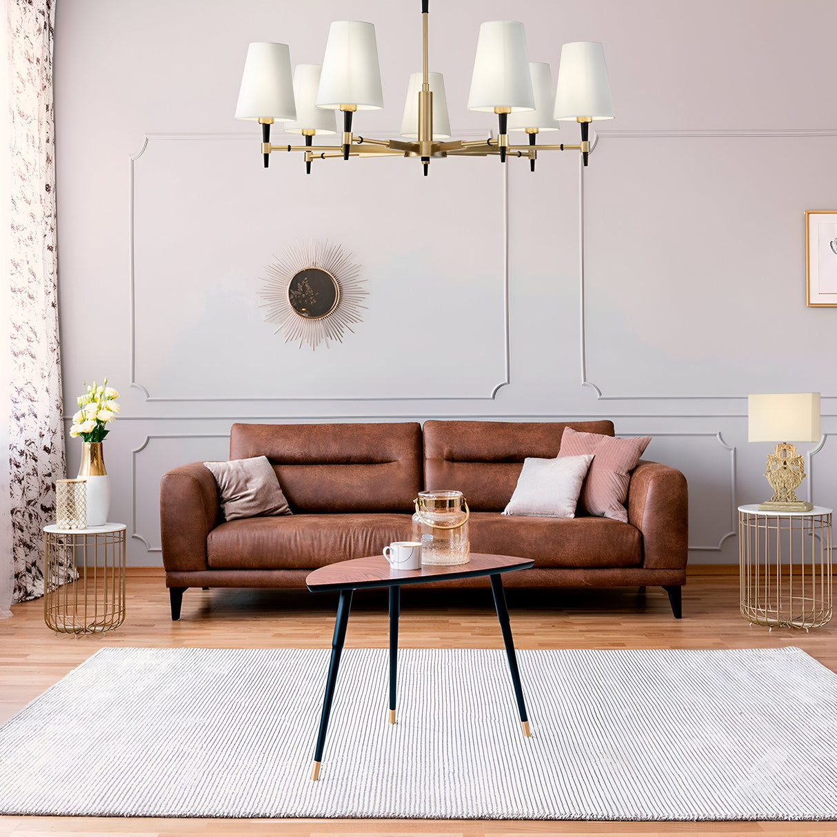 A modern living room features a brown leather sofa with pillows, a wooden coffee table on a light rug, and elegant metal side tables with lamps. Overhead, the brass Zaragoza 7 Light Chandelier with white shades illuminates the space alongside a decorative sunburst wall clock.