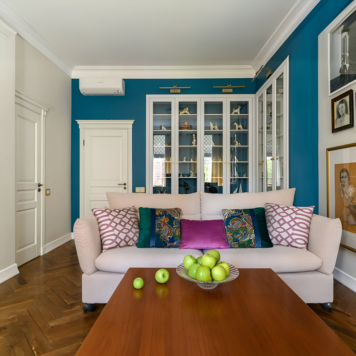 A cozy living room showcases a white sofa with colorful cushions, a wooden coffee table holding green apples, and framed art enhanced by the illumination of a Finelli LED Picture Light. Blue walls, glass cabinets, parquet flooring, and a white door complete the charming atmosphere.