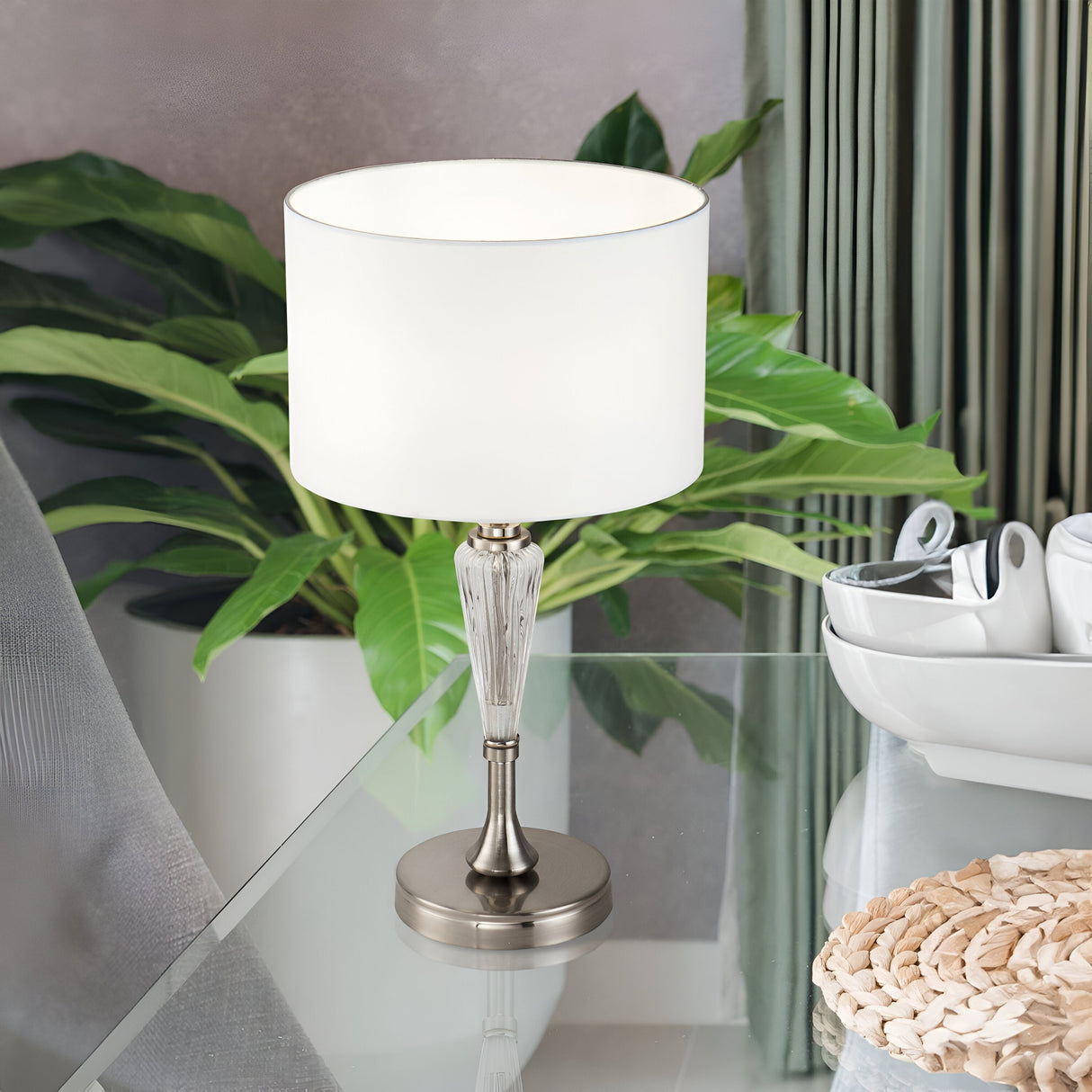 The Alicante Table Lamp - Nickel, featuring a sleek metal base and white cylindrical shade, elegantly rests on a glass table. Alongside it are a potted plant and woven mat near a white bowl with cutlery, set against the vibrant backdrop of a green curtain.