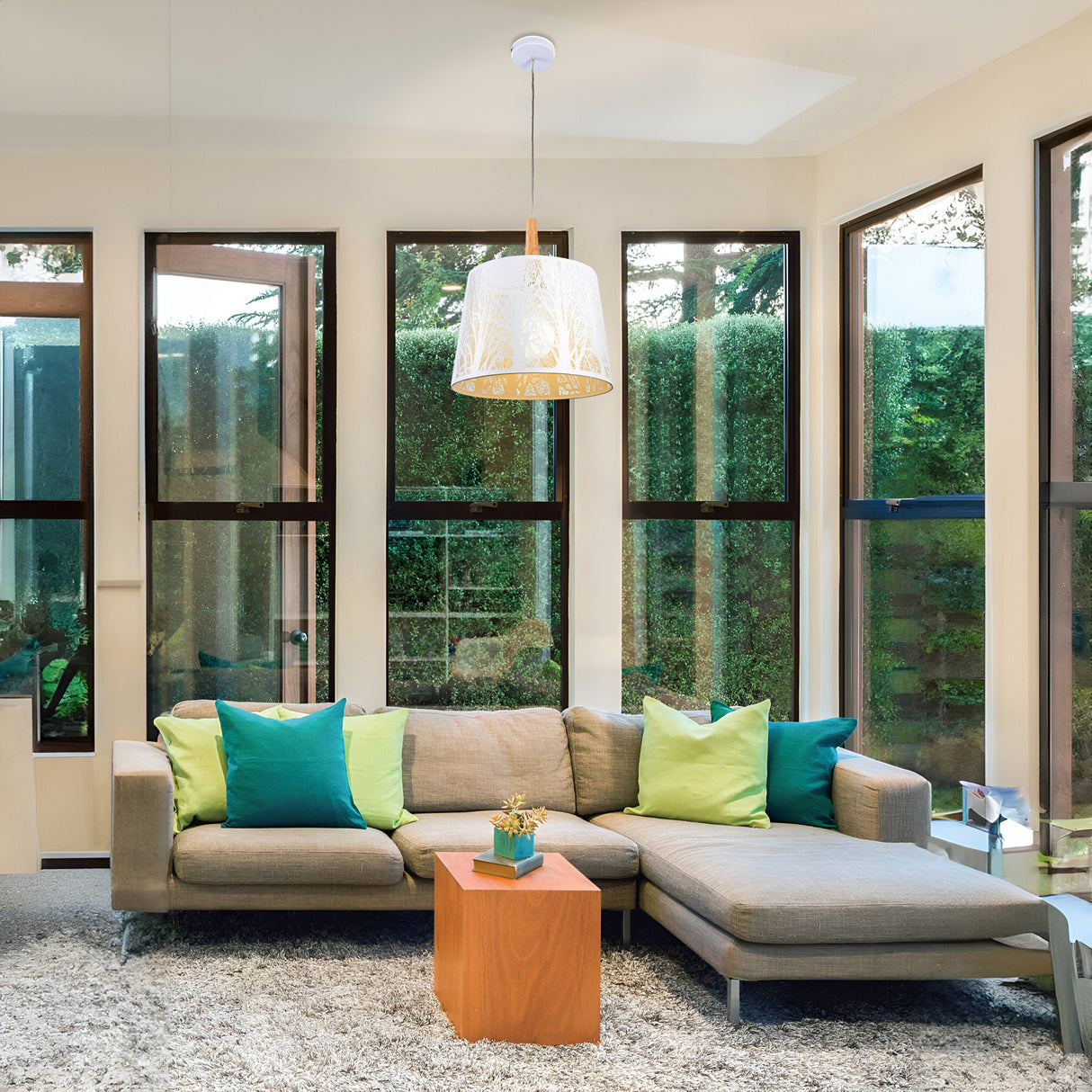 A modern living room with tall windows features a gray sectional sofa accented by blue and green pillows. A small wooden coffee table sits nearby. The Lantern Pendant Light - White With Acid-Etched Glass adds elegance, and lush greenery is visible through the windows.