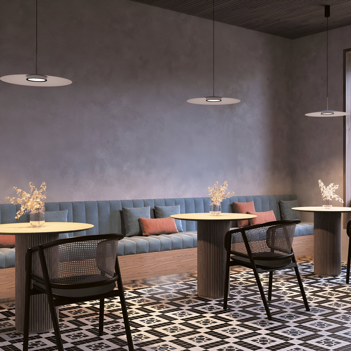 A modern cafe with a minimalist design features a long blue sofa along a grey wall, black and white geometric floor tiles, round tables with decorative vases, black chairs, and the El LED Round Tablet Pendant Light elegantly hanging from the ceiling in its energy-efficient lighting setup.