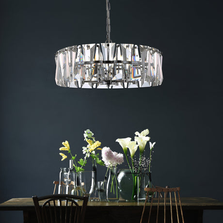 The Puntes 6 Light Chandelier - Chrome elegantly hangs over a wooden dining table. Adorned with vases of lilies and hydrangeas and set against a dark wall, it embodies modern design.