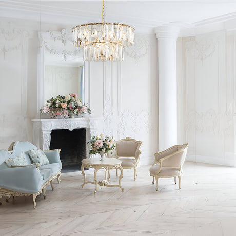 A beautifully elegant room with ornate furniture, featuring the Puntes 12 Light Chandelier in gold. A large mirror sits above a white fireplace adorned with pink and white flowers. Includes a pastel blue couch and cream chairs around a table, creating a classic, luxurious ambiance.