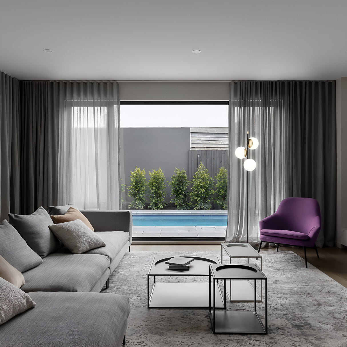 A modern living room boasts a gray sectional sofa, purple armchair, and geometric coffee table exuding vintage charm. Floor-to-ceiling windows overlook a pool and greenery. Gray curtains frame the view, while the Nostalgia Floor Lamp in gold and black adds a minimalist touch overhead.
.